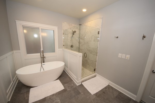 Bathroom with grey tile floor, large white bathtub, and glass shower